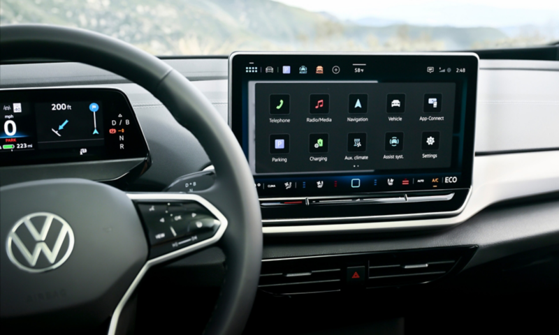 Interior view of a VW showing the steering wheel, dash, and infotainment display with mountains visible through the windshield.