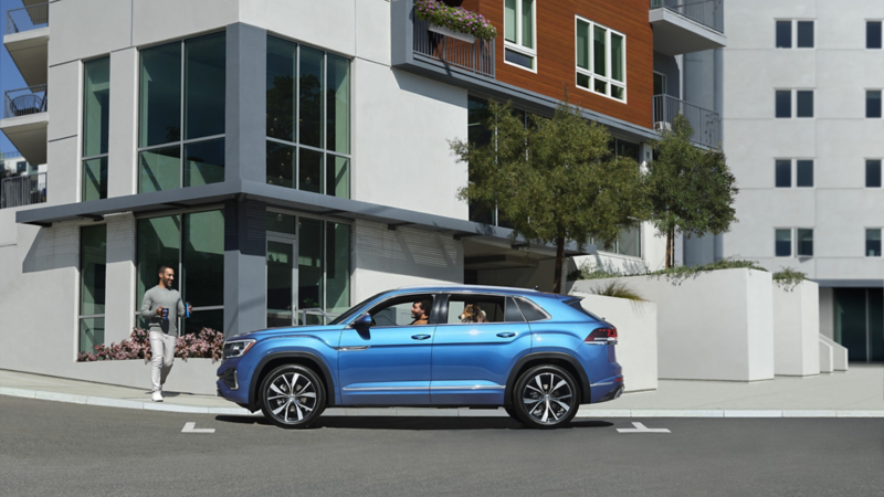 A side profile view of the Atlas Cross Sport shown in Kingfisher Blue Metallic parked in front of a building with a person walking towards it with coffee