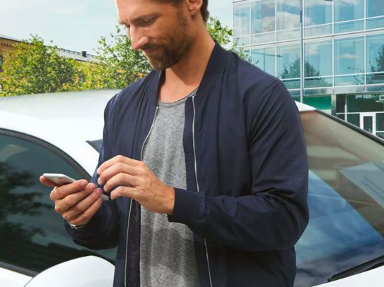 Un hombre a la moda, con una chaqueta ligera, se encuentra en el estacionamiento de un distrito comercial mirando alegremente su teléfono.