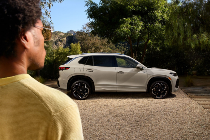 Vista de perfil de un Tiguan en color Sandstone estacionado en un camino bordeado de árboles, visto por encima del hombro de una persona.