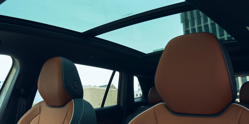 Vista interior del Tiguan en cuero Amber Brown, mirando hacia adelante, con edificios modernos visibles a través de las ventanas y el techo solar panorámico disponible.