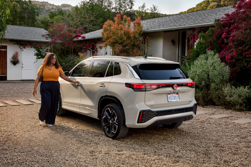 Rear view of a Tiguan shown in Sandstone parked in front of a house while a woman walks toward the driver's door.