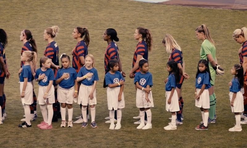 A group of women soccer players line up on a field with young girls standing next to them wearing blue Volkswagen tee shirts.