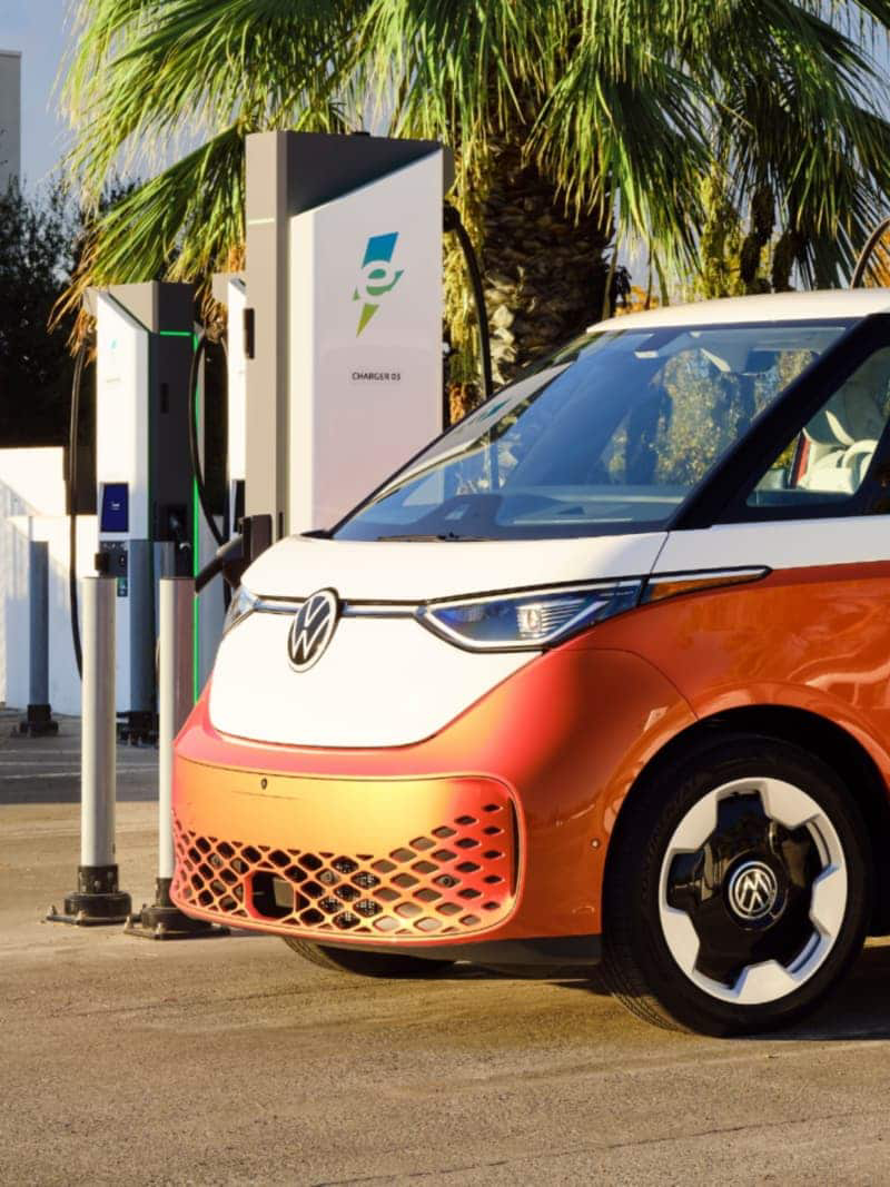 Side view of an ID. Buzz shown in Energetic Orange Metallic and Candy White parked at a charging station with palm trees in the background.