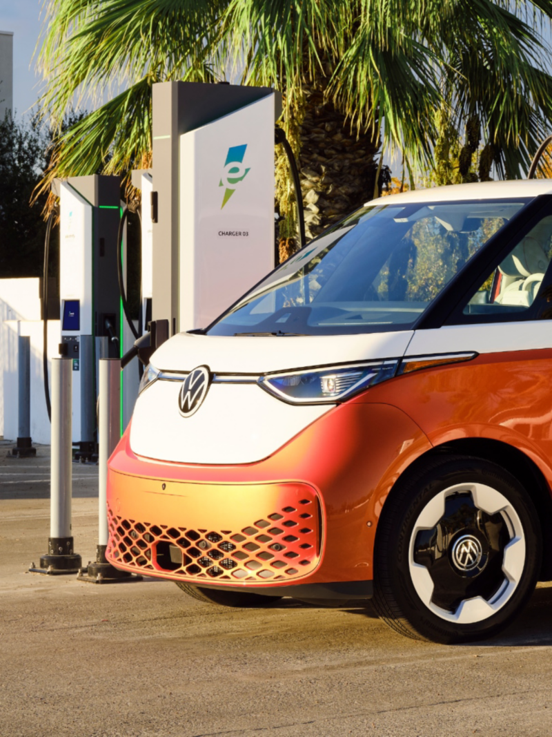 Side view of an ID. Buzz shown in Energetic Orange Metallic and Candy White parked at a charging station with palm trees in the background.