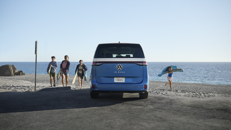 Vista posterior de un ID. Buzz en color Cabana Blue Metallic y Candy White estacionado en una playa con una familia fuera del vehículo llevando equipo de playa y el mar de fondo.