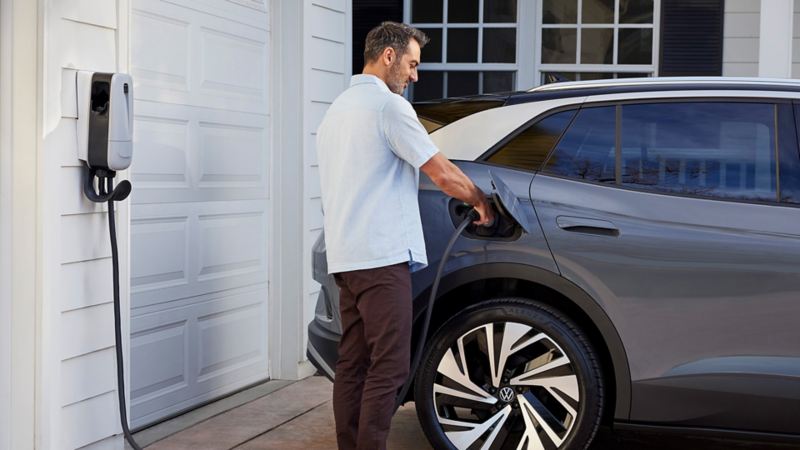 Rear side view of an ID.4 in Pure Gray parked in front of a home garage with a man plugging in the home charger.