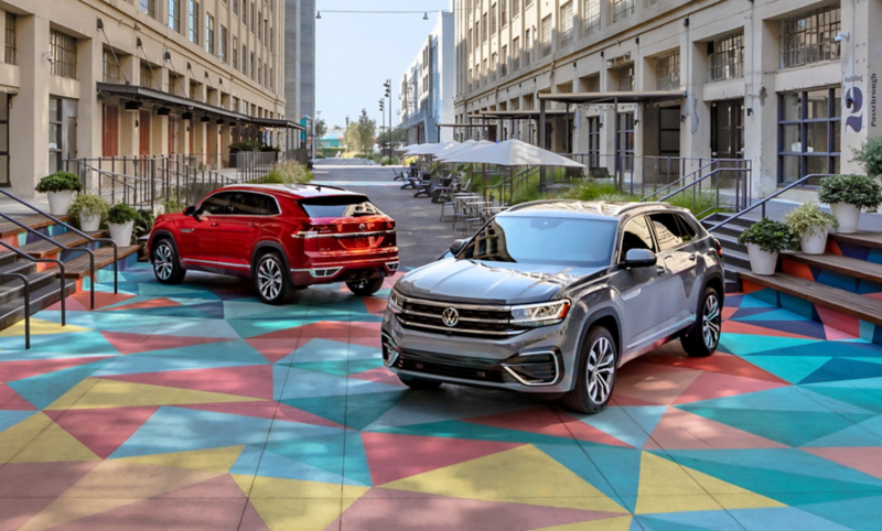 Two Atlas Cross Sports, one in Aurora Red Metallic and one in Pure Gray, parked in a colorful courtyard in between two tall buildings.