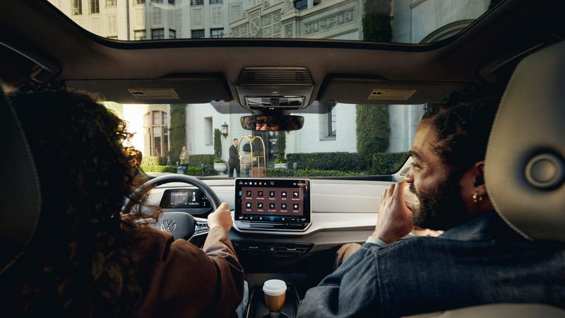 View from inside an ID.4 EV looking out the windshield while a woman drives and a man is smiling in the passenger seat as they approach an upscale hotel.