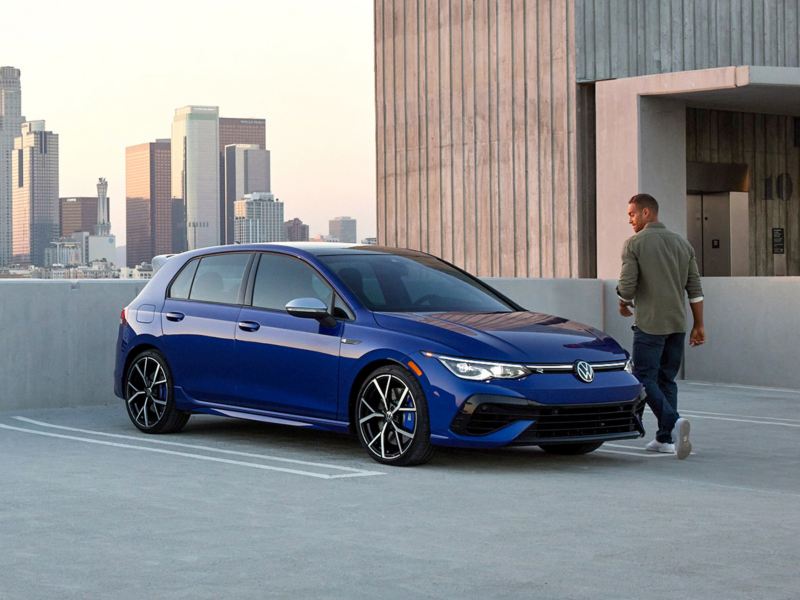 Young man turning to admire a Volkswagen Golf R in Lapiz Blue Metallic parked on rooftop parking lot with city skyline.