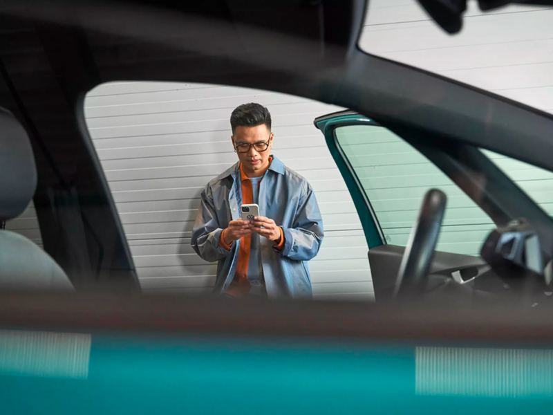Blurred POV from passenger side window of a teal VW with a man leaning against the wall looking at a phone.
