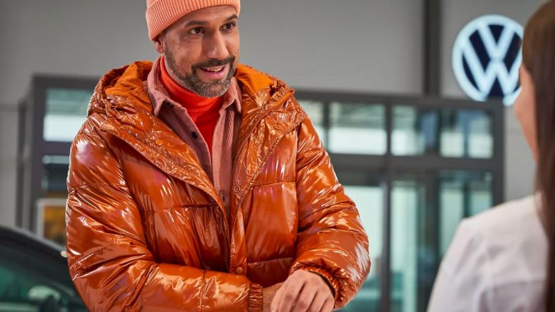 Bearded man with salmon beanie and shiny orange puffer jacket in conversation with woman dealer at VW dealership.
