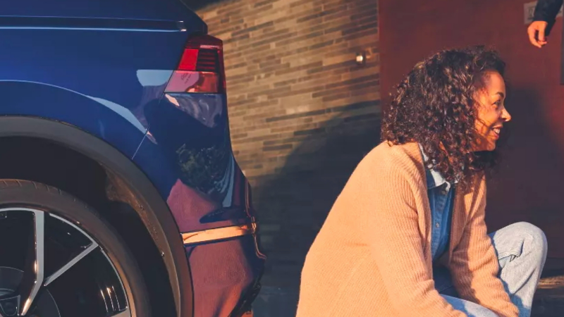 A closeup view of the side rear of a Volkswagen with a woman sitting next to it laughing.