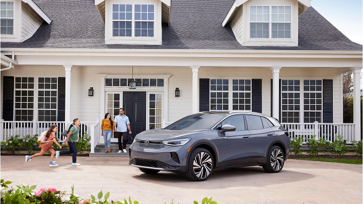2 children are running toward a Gray ID.4 parked in the driveway and two parents are walking out of the front door of a modern home.