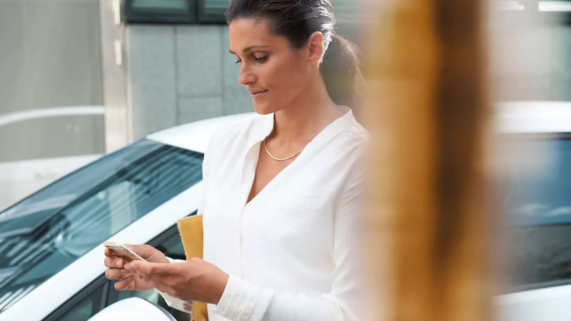 Mujer con cola de caballo baja, collar plateado y blusa blanca mira su teléfono con satisfacción afuera al lado de VW.