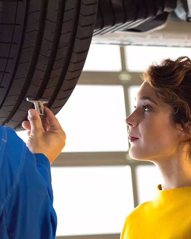 A una joven con un suéter amarillo mostaza se le muestra el estado de la llanta de su VW por parte de un técnico.