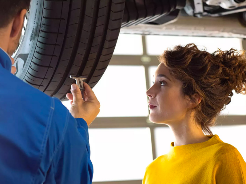 A una joven con un suéter amarillo mostaza se le muestra el estado de la llanta de su VW por parte de un técnico.