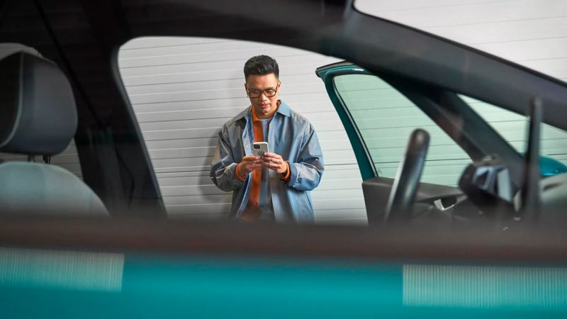 Punto de vista borroso desde la ventana del lado del pasajero de un VW verde azulado con un hombre apoyado contra la pared mirando un teléfono. 