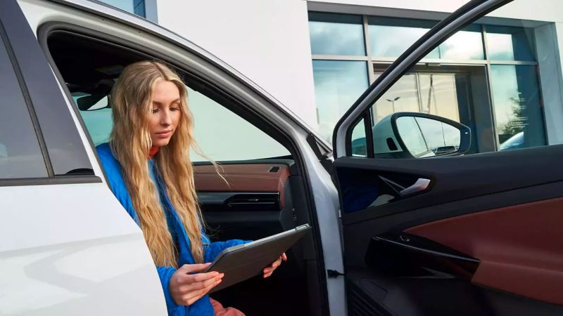 Young woman with long blonde hair sits in passenger seat, door ajar, of white Volkswagen SUV, holding tablet.