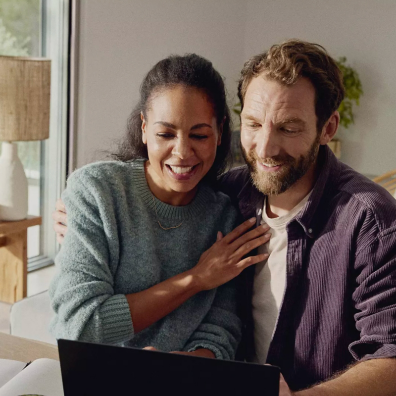 An excited couple with arms wrapped around each other sits in the living room looking at their laptop.