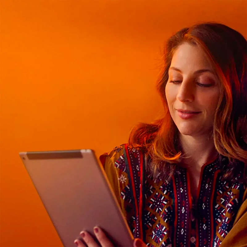Mujer sonriente con blusa bohemia estampada sostiene y mira hacia abajo a la tableta, complementada con un fondo naranja.