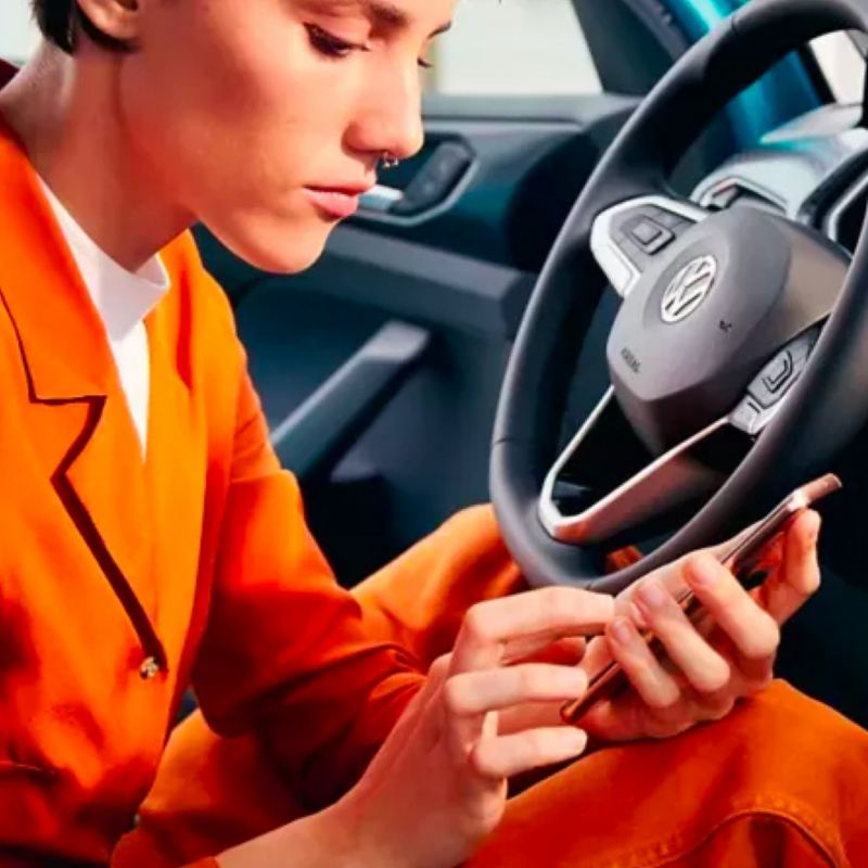 Una joven elegante con un traje de pantalón informal de color naranja quemado se sienta en el asiento del conductor de un VW estacionado que se desplaza por el teléfono.