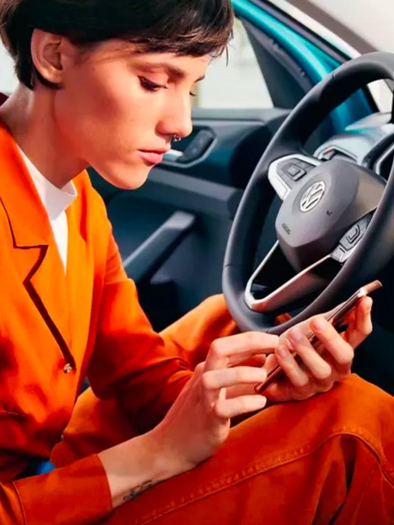 A chic young woman in a casual burnt orange pantsuit sits in the driver’s seat of a parked VW scrolling through phone.