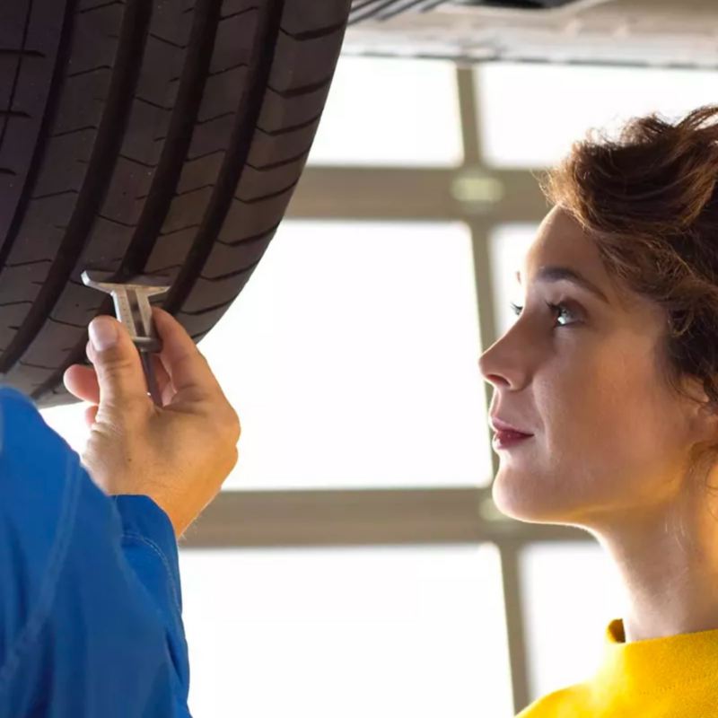 A una joven con un suéter amarillo mostaza se le muestra el estado de la llanta de su VW por parte de un técnico.