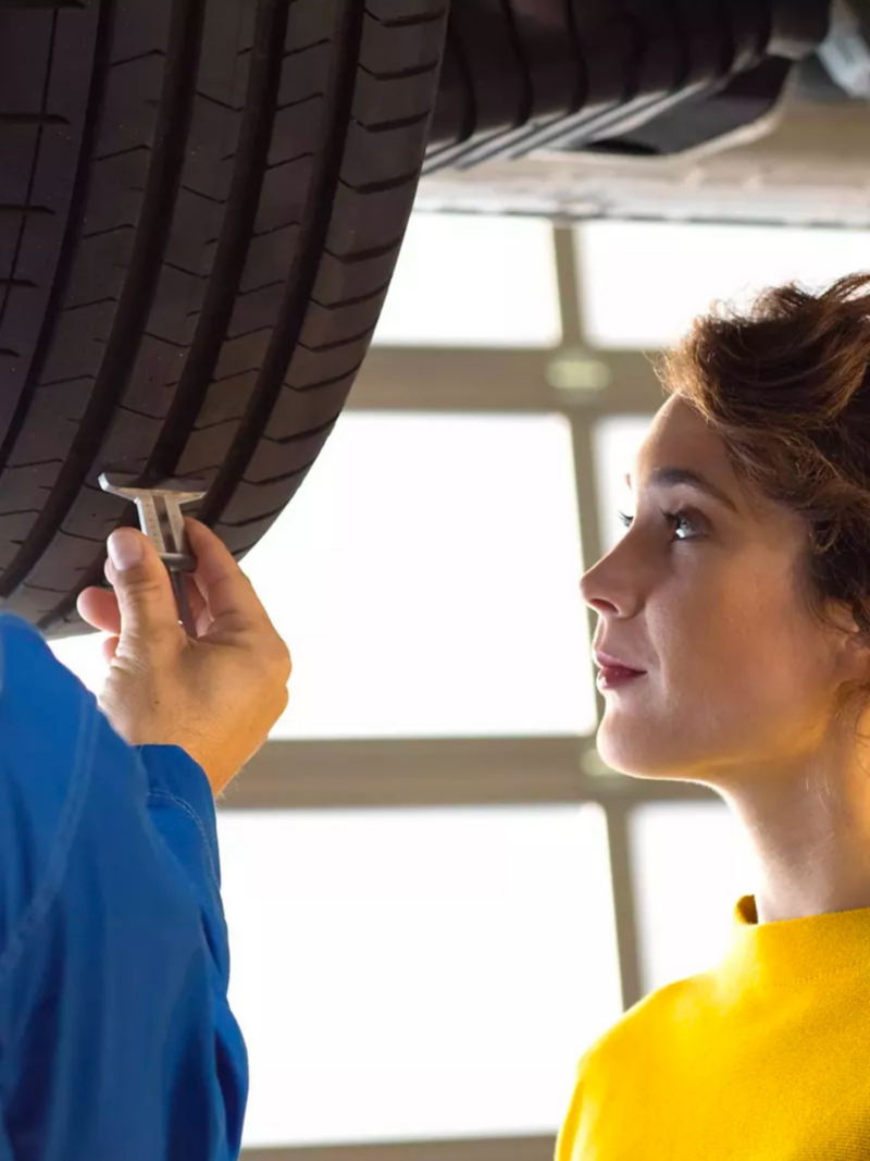 A young woman in a mustard yellow sweater is being shown the status of her VW’s tire by technician.