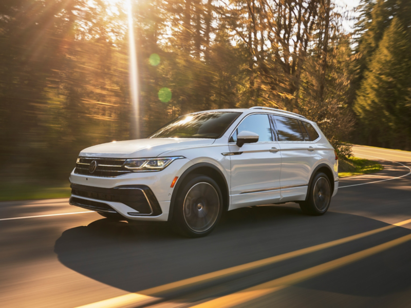 Sunrise shines and bursts through slender trees as a VW Tiguan in Opal White curves around open woodland road.