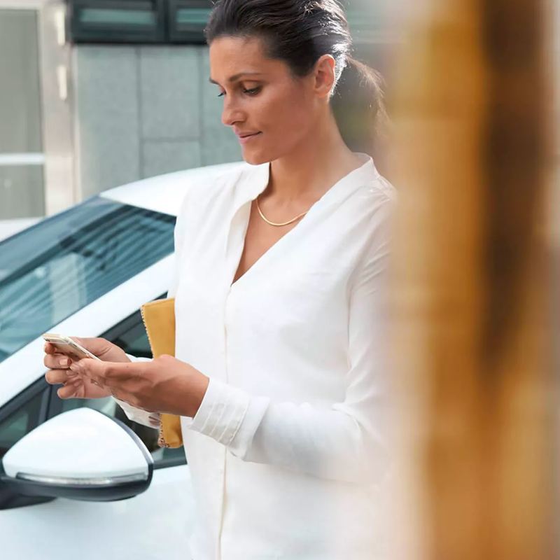 Woman with low ponytail, silver necklace, and white blouse looks down at her phone contently outside next to a white Volkswagen. 