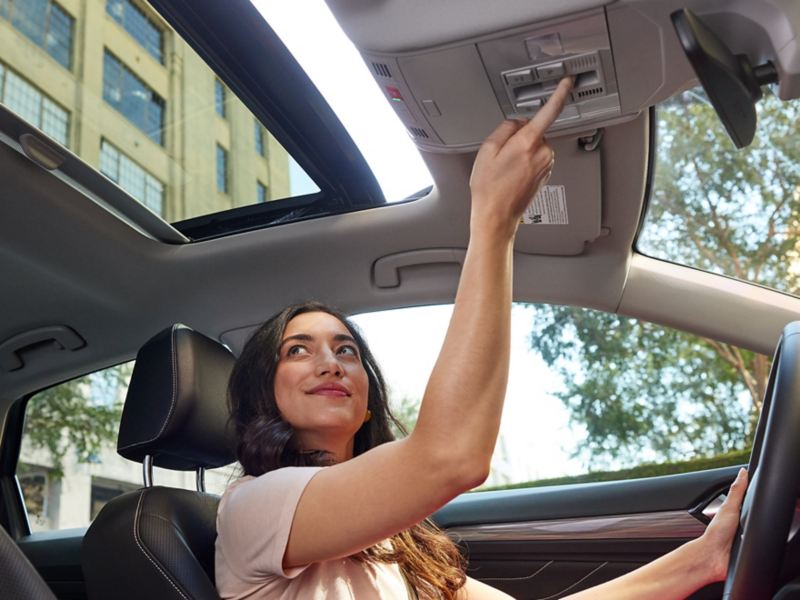 Smiling woman in driver’s seat looks up at opening moonroof of VW, pressing the open button.