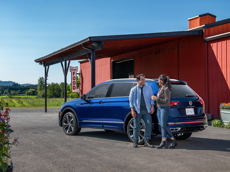 Smiling couple stands next to parked Atlantic blue metallic VW Tiguan SEL R-Line at western off-highway food stop.