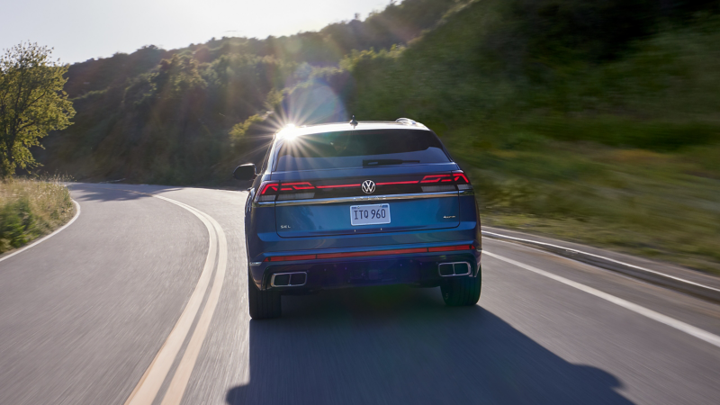 Rear view of a VW Atlas Cross Sport in Kingfisher Blue Metallic driving on a road with grass and trees in the background.