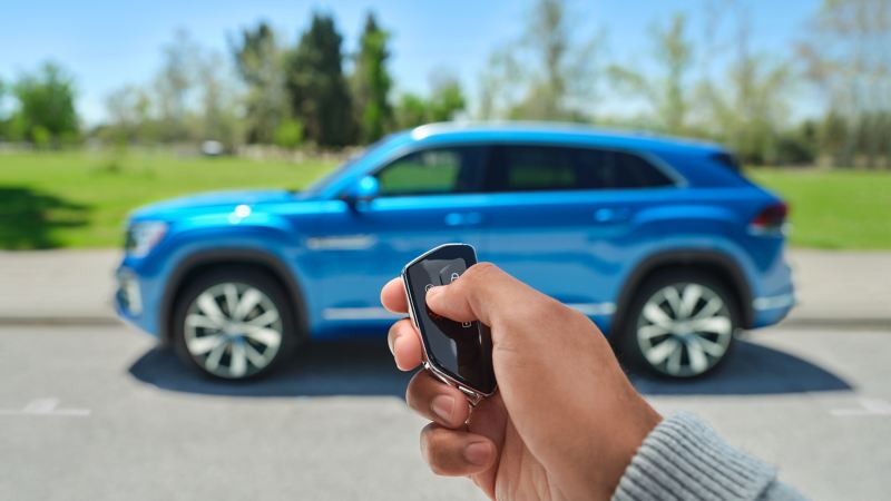 Key fob in front of VW Atlas Cross Sport in Kingfisher Blue Metallic parked along sidewalk with green grass, trees and sunny skies.