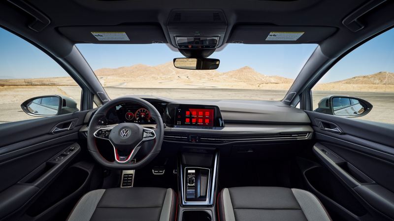 Dashboard view of VW GTI showcases flat-bottom steering wheel, and red-accented, sporty ambiance, overlooking sunny desert.