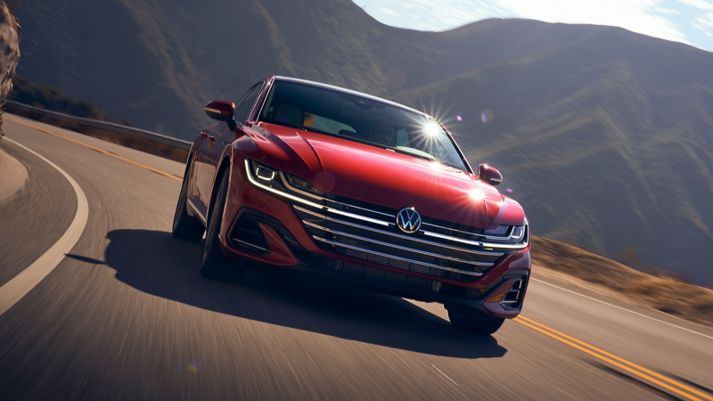 Head on view of VW Areton in Kings Red Metallic winding along road, backgrounded by mossy mountains and blue skies.