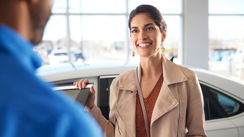 A woman smiles standing next to a white VW at a Volkswagen dealership.