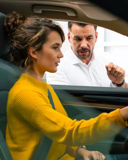 Toma de una joven con un suéter color amarillo mostaza sentada en el asiento del conductor de una Tiguan de Volkswagen con el gerente de ventas.
