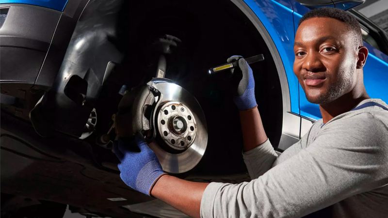 Worker in service garage smiles with flashlight in-hand, cradling the exposed brake rotor of a suspended VW.