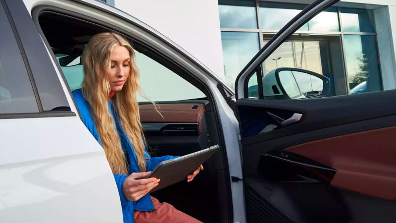 Young woman with long blonde hair sits at passenger’s seat, door ajar, of white Volkswagen SUV, holding tablet.