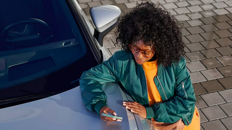 Woman in silk teal bomber jacket, sweatshirt dress, stylish glasses, and matching fanny pack texting and leaning against VW.