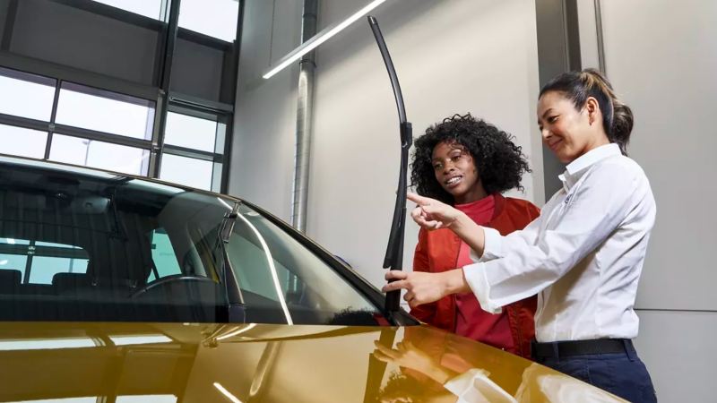Woman dealer in service garage holds up windshield wiper of VW Arteon in Kurkuma Yellow to woman customer.