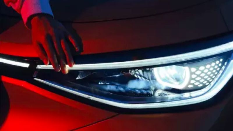 An up-close view of the illuminated headlight of a VW ID.4 in Aurora Red Metallic with a hand on hood.