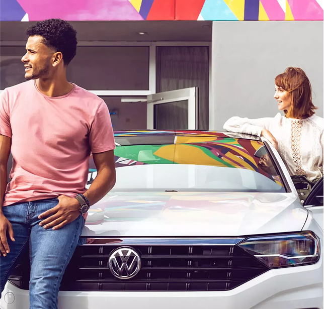 A man in a t-shirt sits on a Volkswagen looking to the left and a woman is standing at behind the driver door that is open in front of a VW dealership