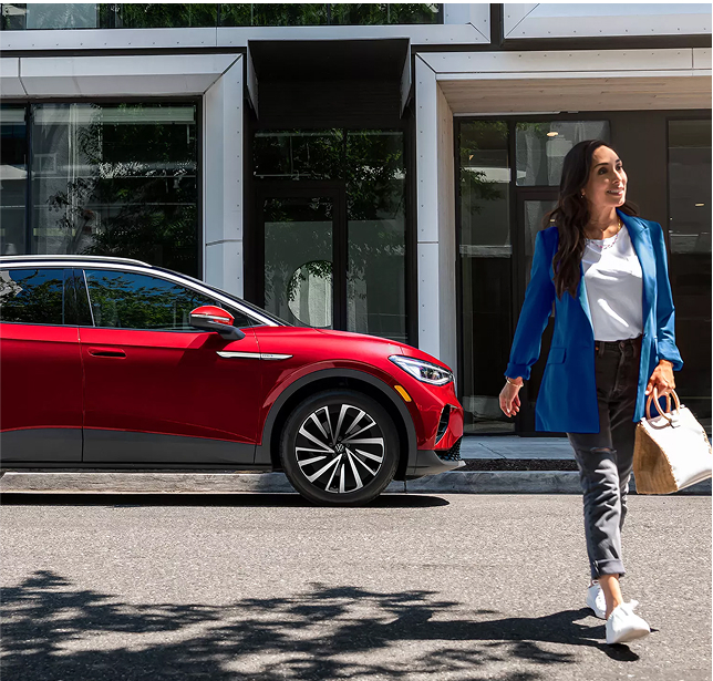 A woman walks away from an Aurora Red Metallic ID.4 SUV that is parked on an urban street in front of a modern office building.