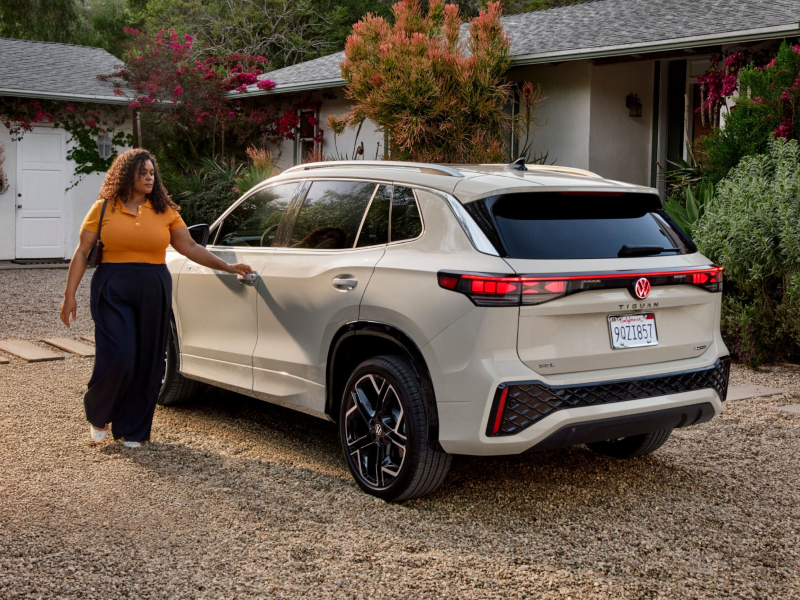 Rear view of a Tiguan shown in Sandstone parked in front of a house while a woman walks toward the driver's door.