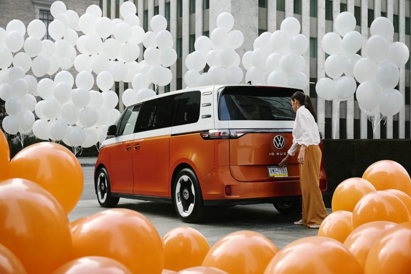 Rear view of a person opening the liftgate of an ID. Buzz shown in Energetic Orange Metallic and Candy White parked in a a modern setting with orange and white balloons around the vehicle.