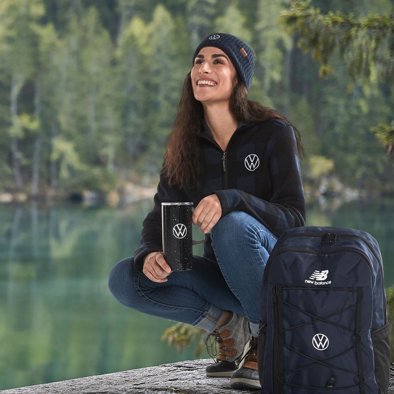 A woman wearing a VW branded fleece is holding a VW branded beverage tumbler with a VW branded backpack next to her and is couching on a rock with a lake and forest in the background.