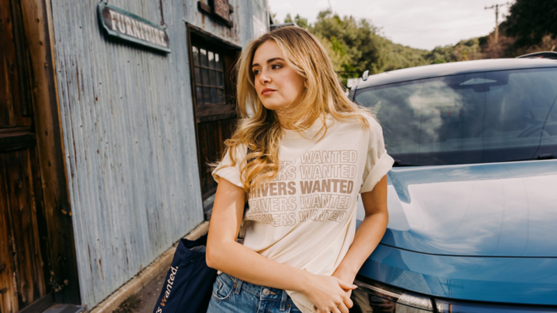 A woman leaning on a VW wearing Drivers wanted merch in front of rustic furniture store with trees in the background.